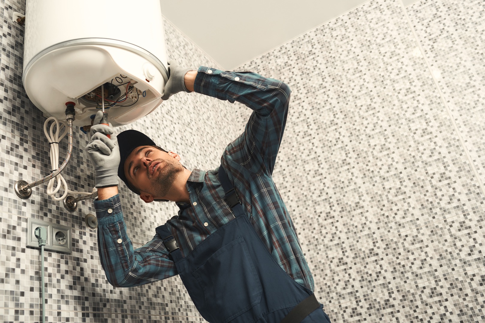 Portrait of young handyman in uniform setting up electric heating boiler at home. Horizontal shot