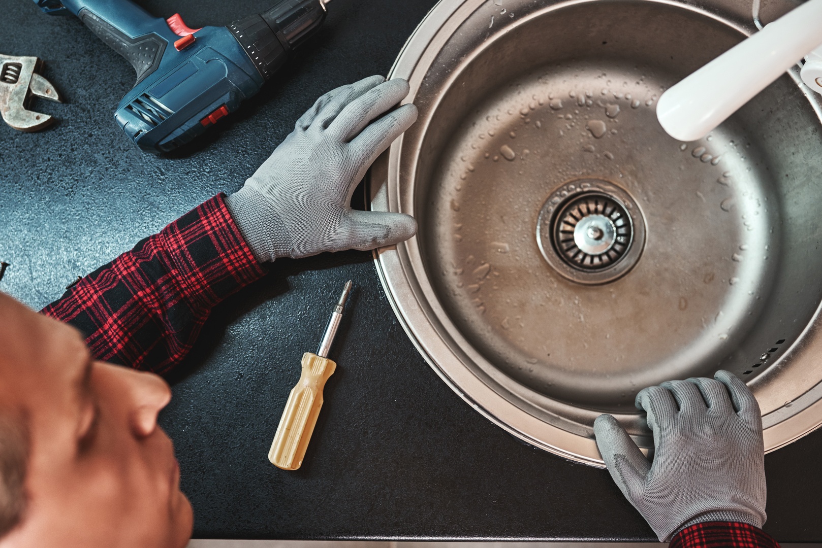 Inside view. Close-up of handsome plumber repairing sink in kitchen. Photo of metalic sink and handyman with screwdriver